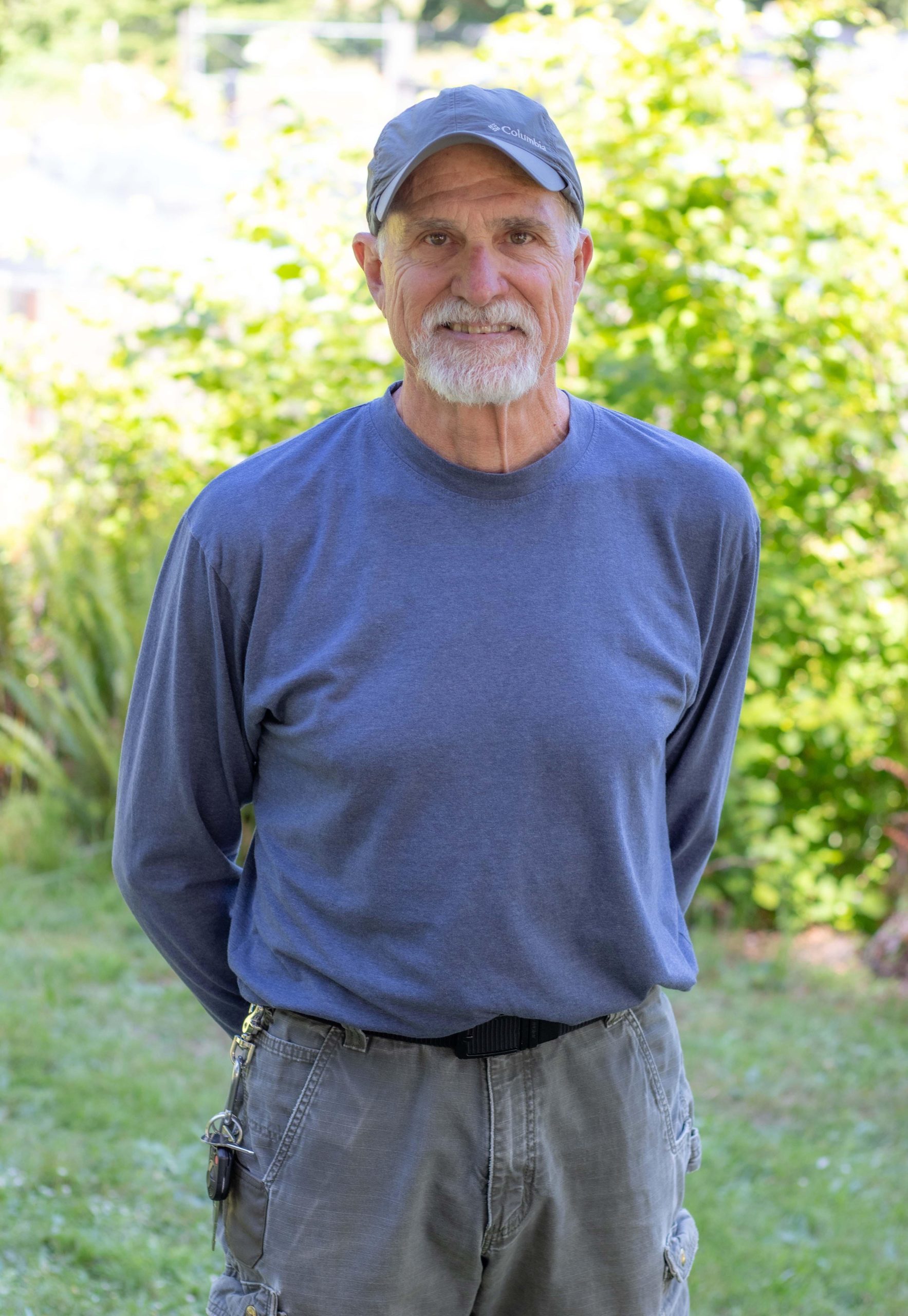 Dennis Quenneville for Yamhill SWCD Website Dennis Quenneville, a man with a mustache and beard, wearing a hat and standing front of a blurred green plant background.